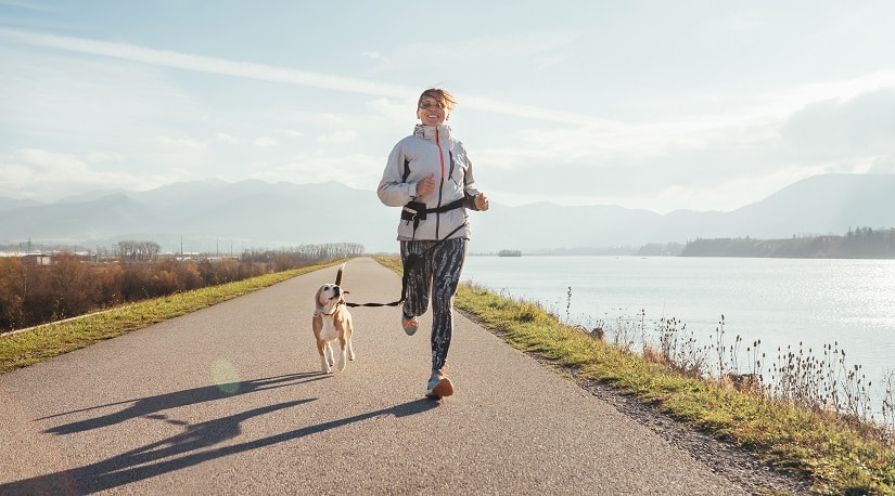 Eine Frau joggt mit ihrem Hund an einem Fluss und trägt dabei einen Jogginggurt für Hunde
